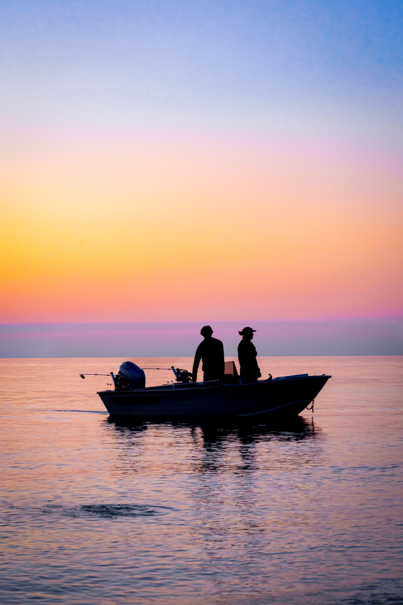A boat with two silhouetted passengers crossing gentle water beneath a pastel sunset